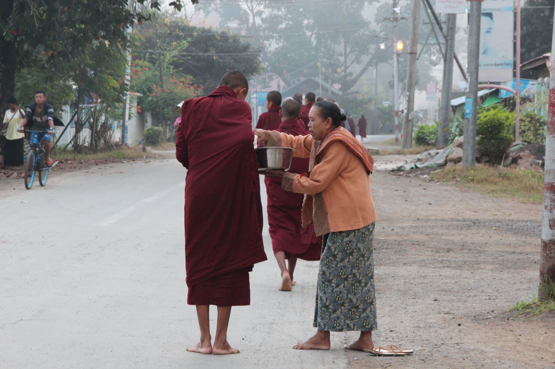 คนยืนถือถุงอาหารรอตักบาตรพระสงฆ์บนทางเท้าตอนเช้า