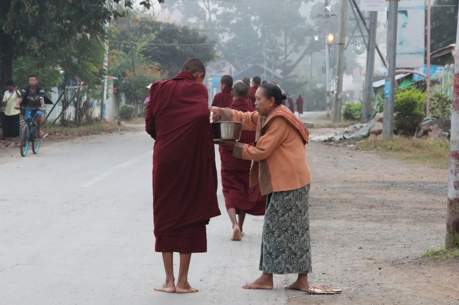 คนยืนถือถุงอาหารรอตักบาตรพระสงฆ์บนทางเท้าตอนเช้า
