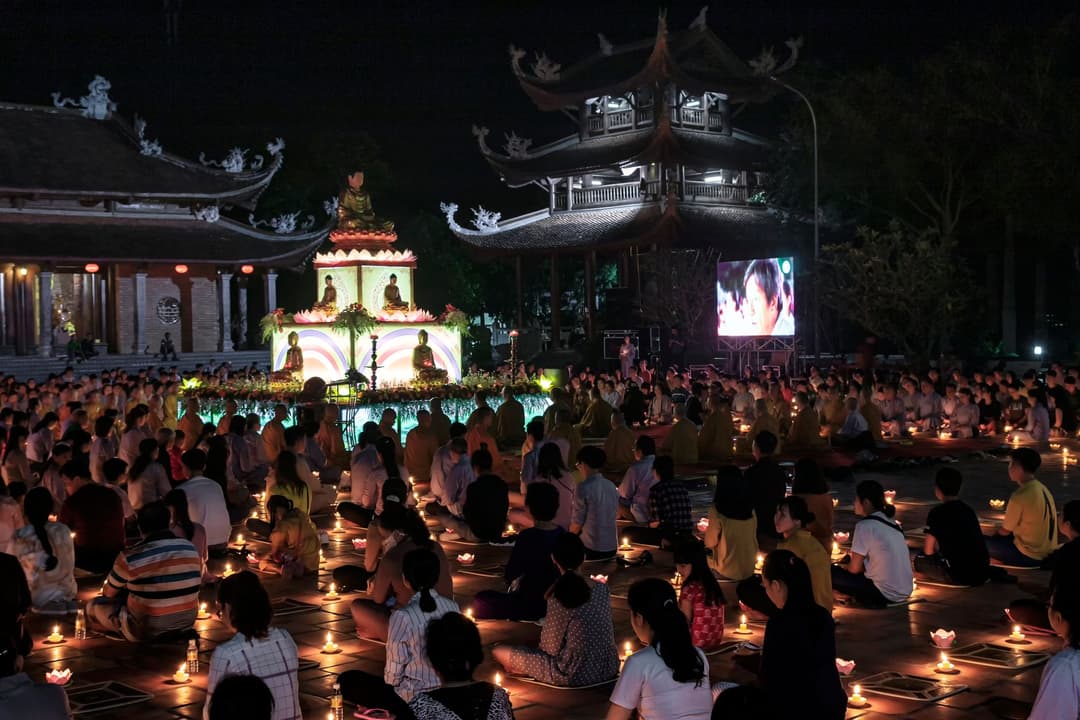 คนจำนวนมากนั่งขัดสมาธิถือเทียนจุดไฟในลานวัดยามค่ำคืน มีพระพุทธรูปสีทองและเจดีย์เป็นฉากหลัง พร้อมจอภาพขนาดใหญ่ด้านข้าง