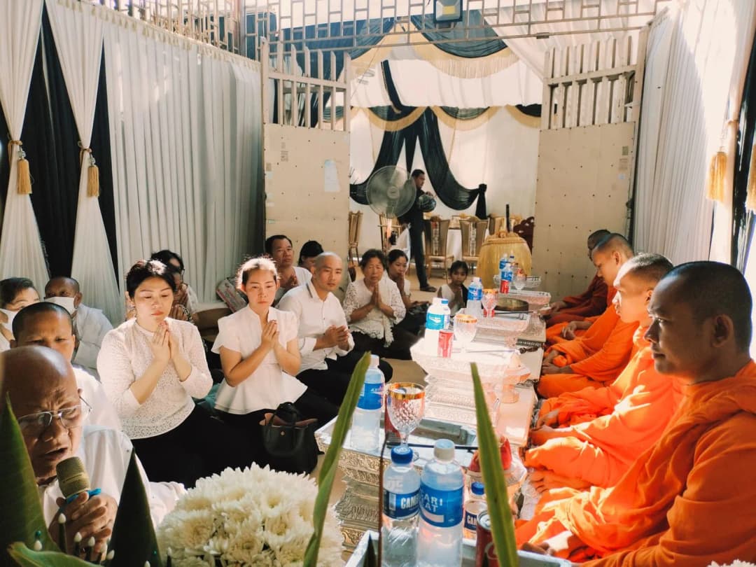 พระสงฆ์นั่งสวดมนต์ในบ้านขณะเจ้าของบ้านนั่งพนมมือฟังอยู่ใกล้ ๆ