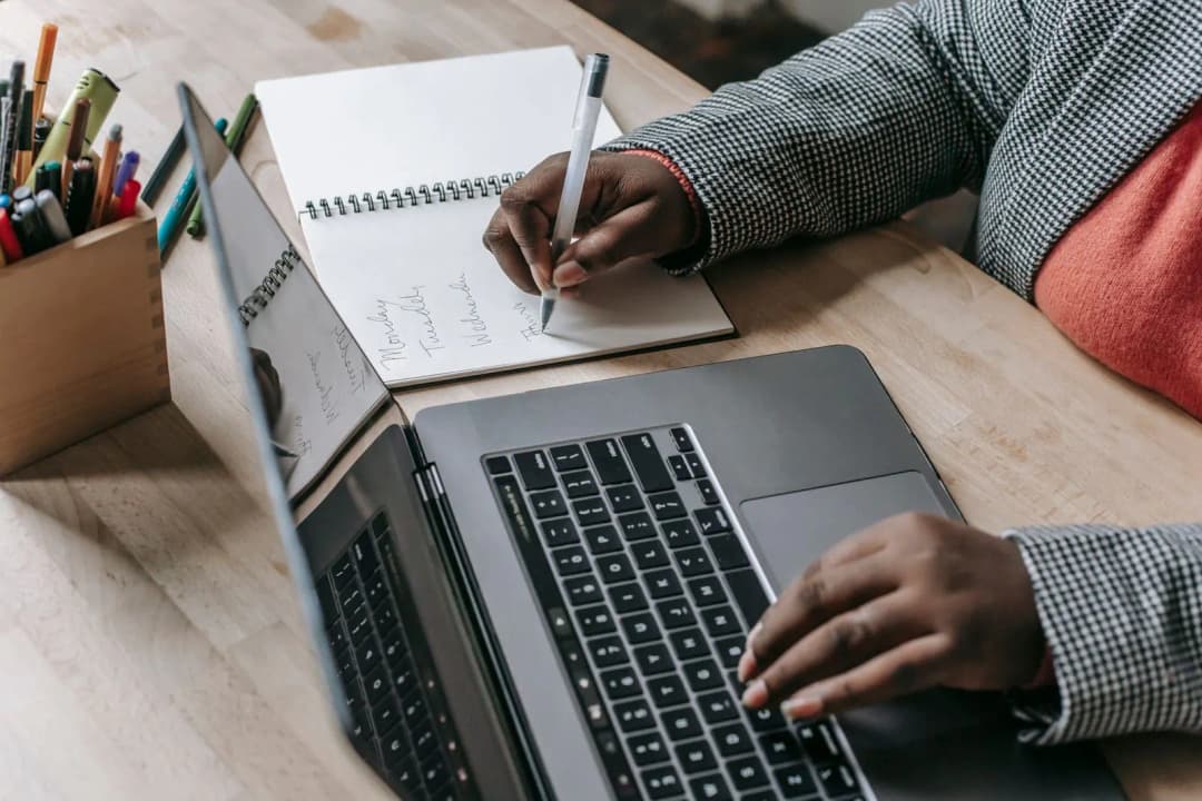 มือกำลังจดบันทึกวันจันทร์ถึงวันพฤหัสในสมุดข้างแล็ปท็อปสีเงินที่เปิดอยู่บนโต๊ะไม้ สื่อถึงการวางแผนเริ่มธุรกิจออนไลน์ทีละขั้น