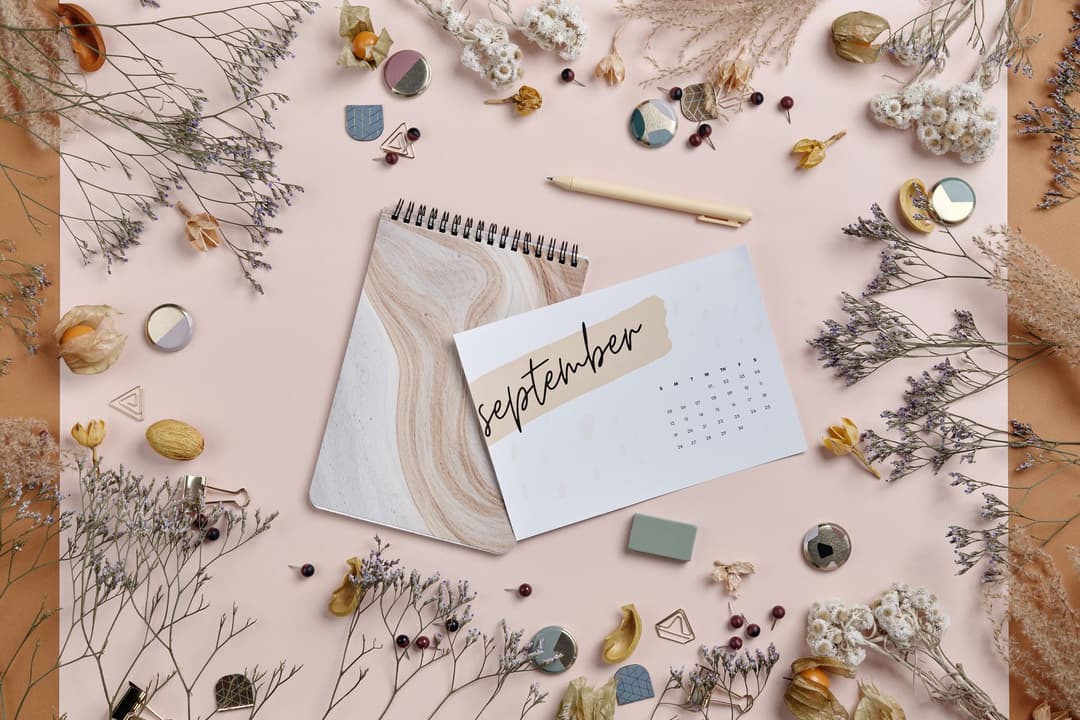 การ์ดปฏิทินเขียนคำว่า September วางอยู่บนสมุดโน้ตลายหินอ่อน ล้อมรอบด้วยดอกไม้แห้งและอุปกรณ์เครื่องเขียนสีพาสเทล